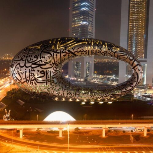 Night cityscape with a large circular Arabic calligraphy sculpture atop a hill by a freeway in Dubai's skyline.