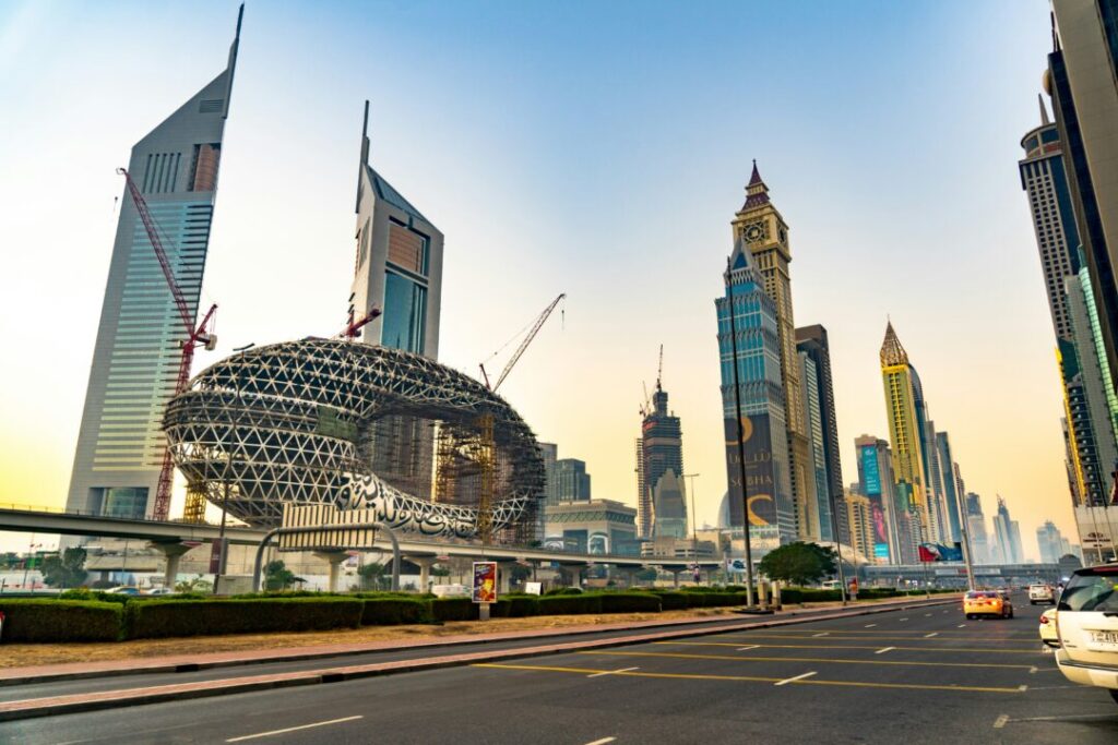 Skyline of Shanghai's Lujiazui district at sunset, featuring a circular lattice building under construction and tall skyscrapers along a wide roadway.