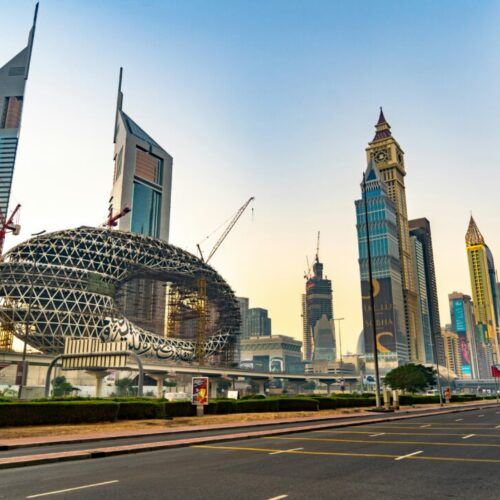 Skyline of Shanghai's Lujiazui district at sunset, featuring a circular lattice building under construction and tall skyscrapers along a wide roadway.