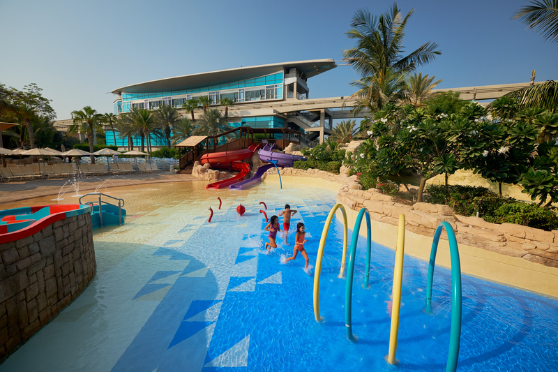 Children play in a shallow bright-blue pool at a tropical water park with colorful slides and a modern building behind.