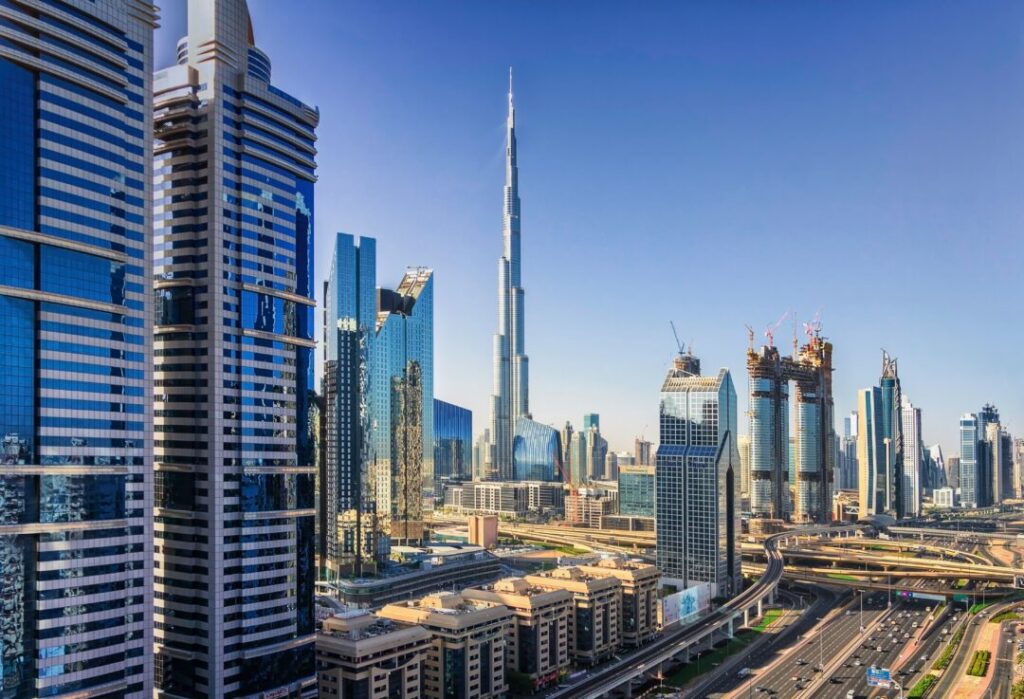 Dubai skyline with Burj Khalifa rising among glass skyscrapers and a multi-lane highway in the foreground, under a clear blue sky.