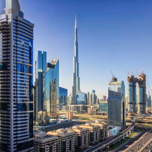 Dubai skyline with Burj Khalifa rising among glass skyscrapers and a multi-lane highway in the foreground, under a clear blue sky.