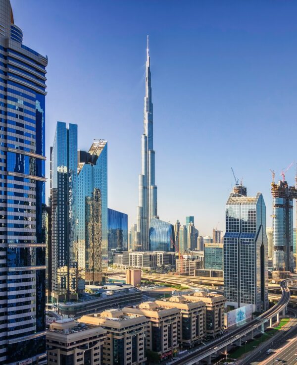 Dubai skyline with Burj Khalifa rising among glass skyscrapers and a multi-lane highway in the foreground, under a clear blue sky.