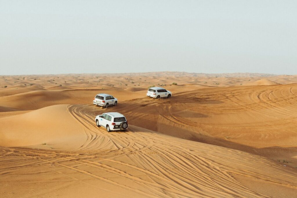 Three white SUVs drive across golden sand dunes in a desert landscape, leaving tire tracks behind them.