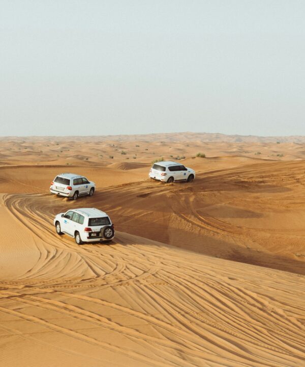 Three white SUVs drive across golden sand dunes in a desert landscape, leaving tire tracks behind them.