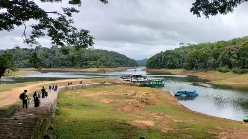People walk along a stone jetty next to a calm river with boats docked by a forested shoreline behind them.