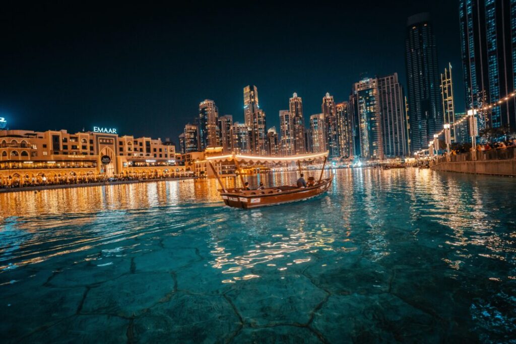 Night view of a illuminated harbor with a traditional wooden boat sailing on turquoise water and towering skyscrapers in the background (Dubai Marina).