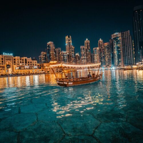 Night view of a illuminated harbor with a traditional wooden boat sailing on turquoise water and towering skyscrapers in the background (Dubai Marina).