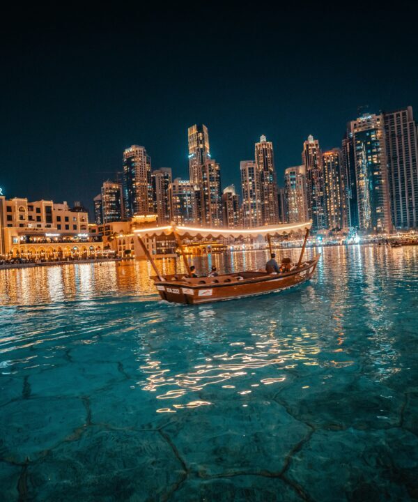 Night view of a illuminated harbor with a traditional wooden boat sailing on turquoise water and towering skyscrapers in the background (Dubai Marina).