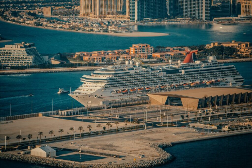 Cruise ship Carnival Splendor docked at a marina with a city skyline and palm-lined promenade in the background.