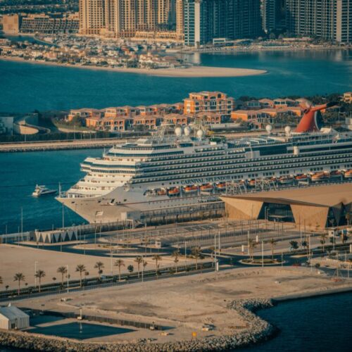 Cruise ship Carnival Splendor docked at a marina with a city skyline and palm-lined promenade in the background.