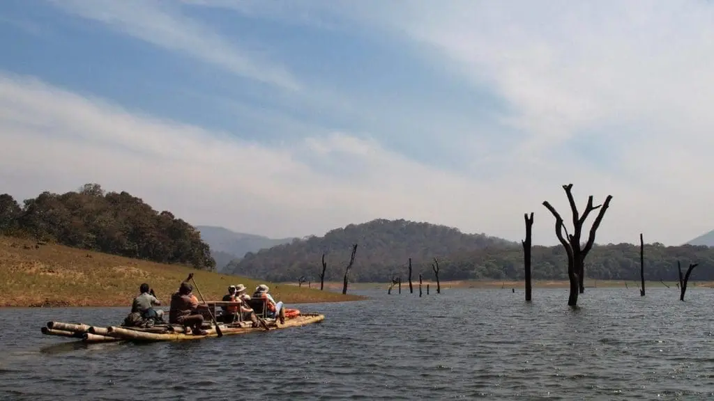 A group of people paddling a makeshift raft across a calm lake, with bare dead trees standing in the water and distant hills under a blue sky.