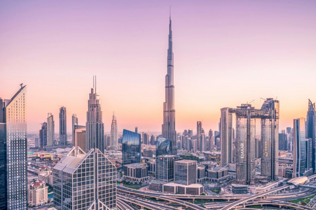 Dubai skyline at sunset with the Burj Khalifa towering above a cluster of skyscrapers and foreground highways.