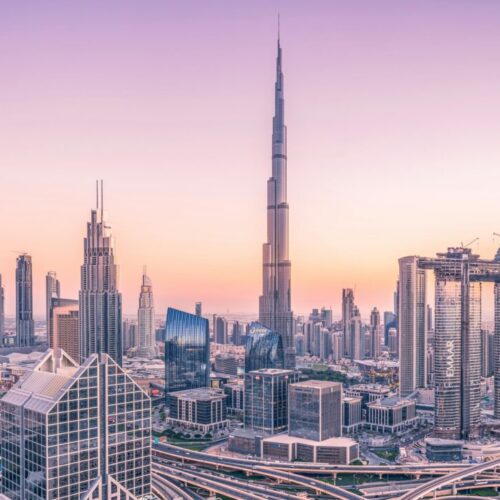 Dubai skyline at sunset with the Burj Khalifa towering above a cluster of skyscrapers and foreground highways.