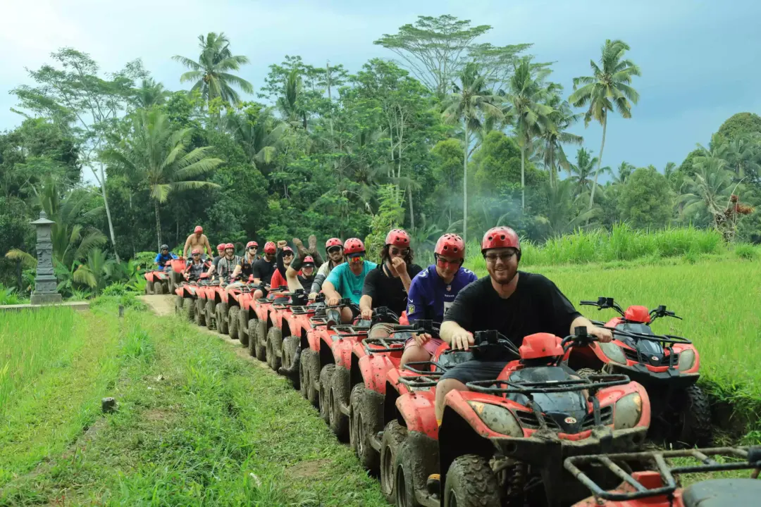 Group of people wearing red helmets riding red ATVs in a muddy path through a lush tropical field with palm trees behind them.