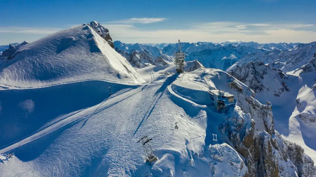 Snow-covered alpine ridge with ski-lift towers and cables under a clear blue sky, mountain range in the distance (informative).