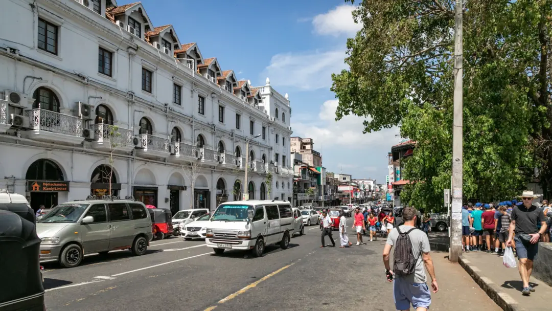 Busy city street with a white colonial building featuring arches and balconies on the left, cars parked and driving, pedestrians crossing. Tall trees on the right under blue sky.