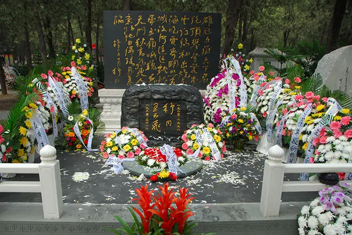 Memorial shrine with a central stone tablet surrounded by colorful flower wreaths at a gravesite or cemetery, with gold Chinese script on a dark backdrop and petals scattered on the ground.