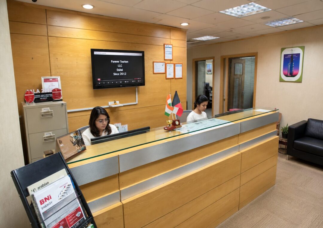 Reception desk with two staff working behind it, Indian and UAE flags on the counter, and a wall-mounted screen showing Forever Tourism LLC Dubai Since 2012.