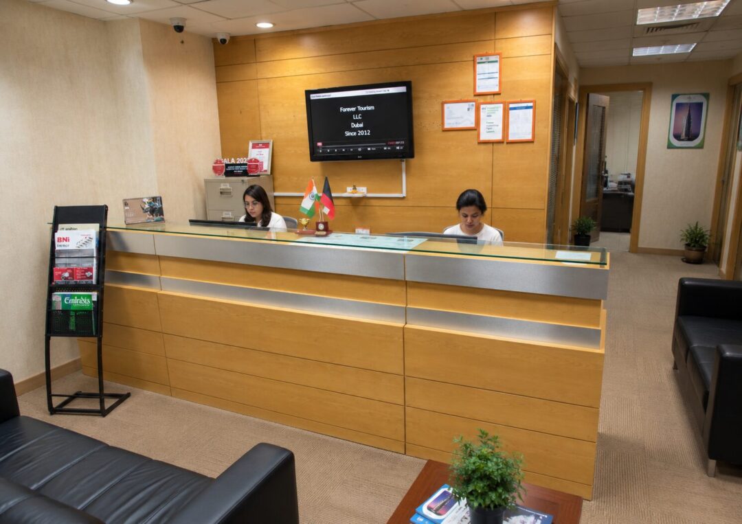 Reception desk in a lobby with two female staff behind a glass counter, Indian flag and another flag on the desk, and a wall-mounted monitor behind them.