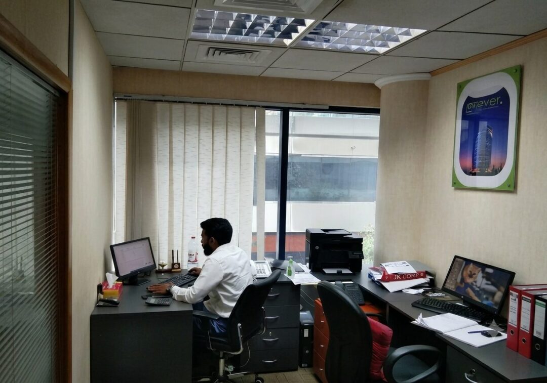 Office worker with a beard sits at a desk typing on a computer near a large window with vertical blinds.
