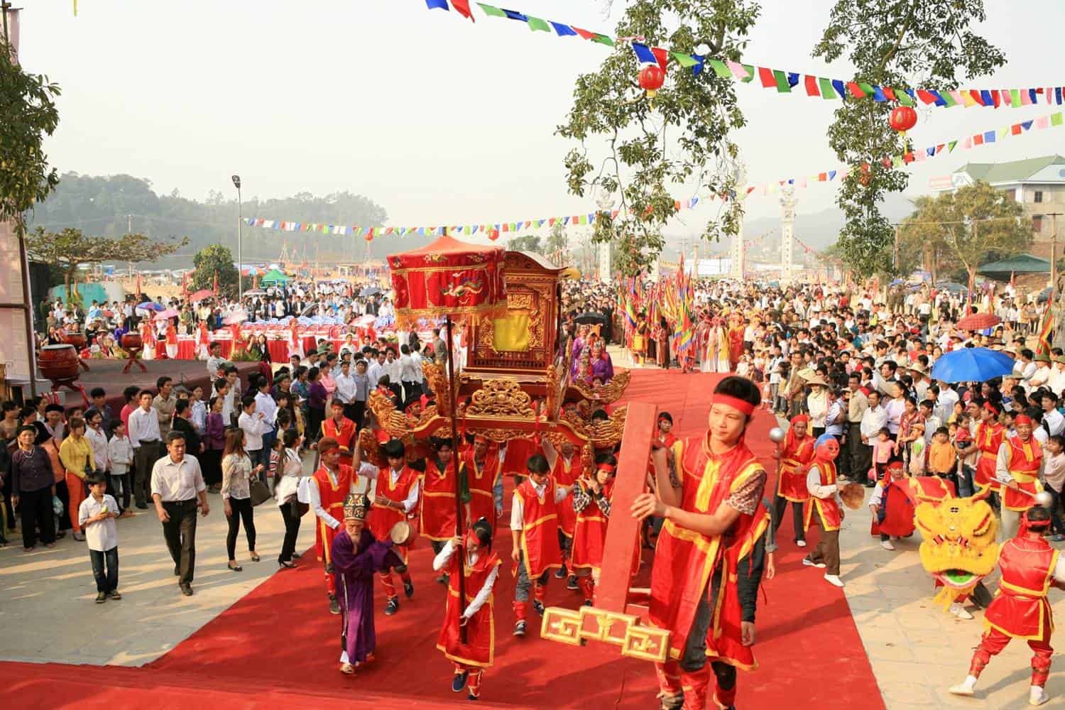 Vibrant outdoor festival with a red-carpet parade, ornate float, and performers in red costumes among a large crowd of spectators.