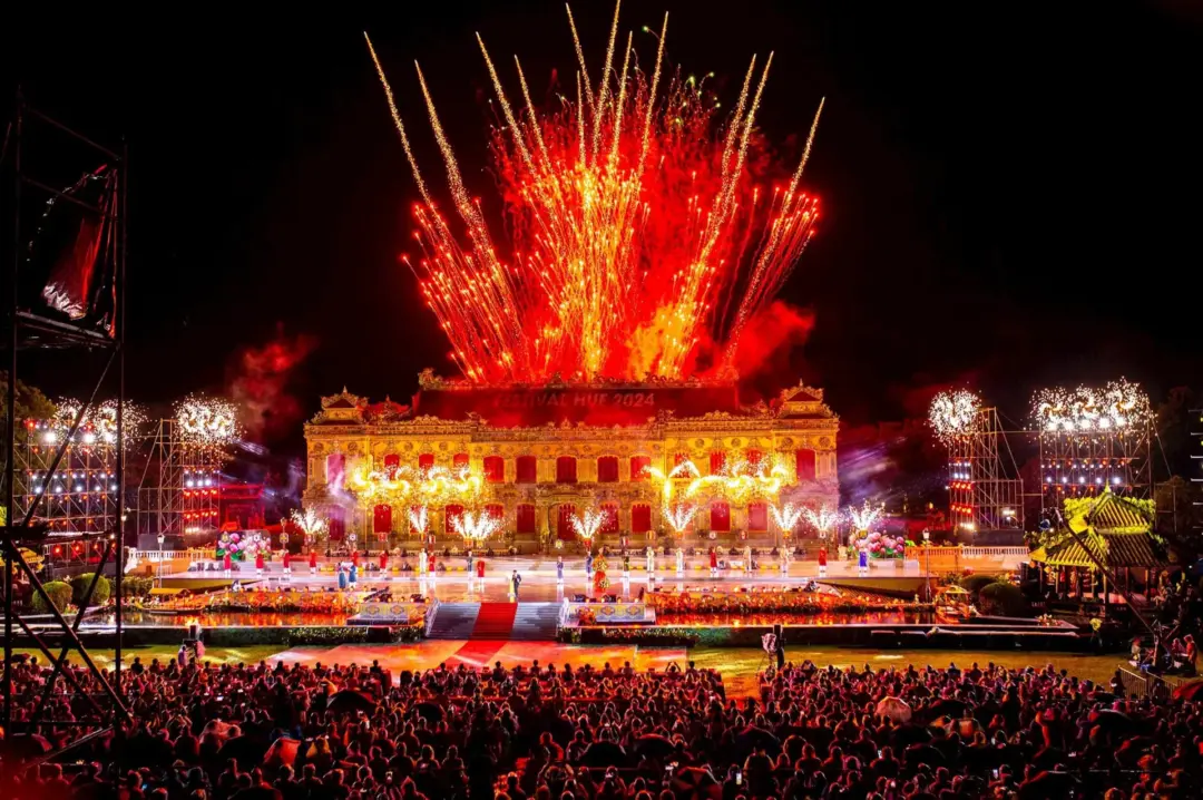 Fireworks explode above a grand illuminated building and a crowded stage audience at night festival.