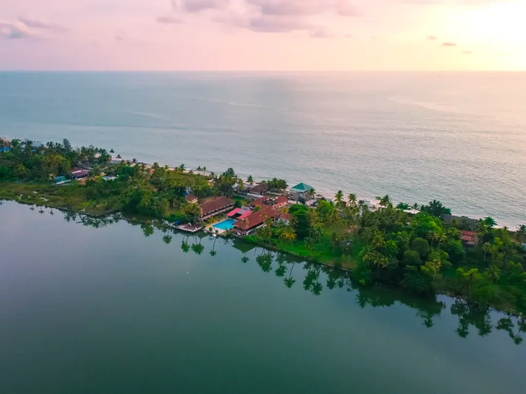 Aerial view of a tropical coastal village with red-roofed houses along a narrow land strip between a calm lagoon and the sea at sunset.