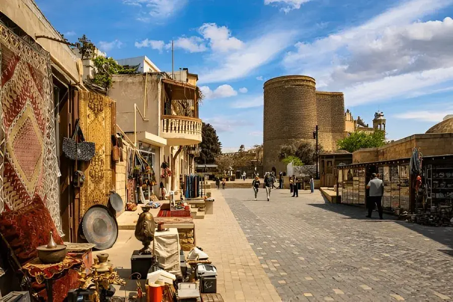 A sunlit cobblestone street in a historic market town with shops displaying rugs, pottery, and textiles along the left, and a stone tower in the distance under a blue sky.