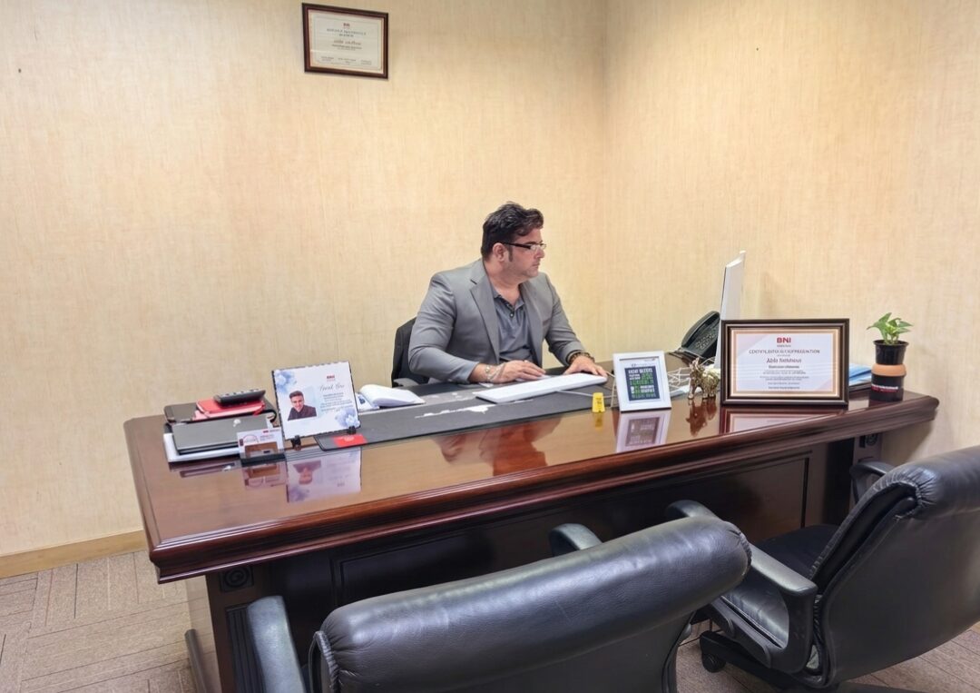 Man in a gray blazer sits at a large polished wooden desk in an office, working with papers under beige walls with framed certificates nearby
