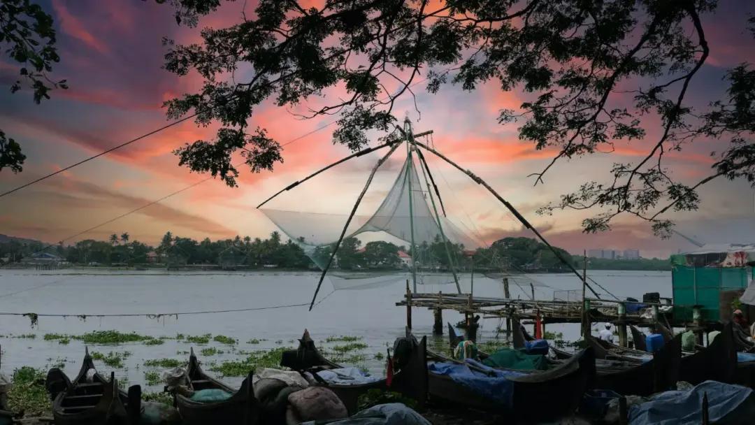 Sunset over a river with a large triangular fishing net rig, boats moored along a wooden jetty, and tree silhouettes overhead.