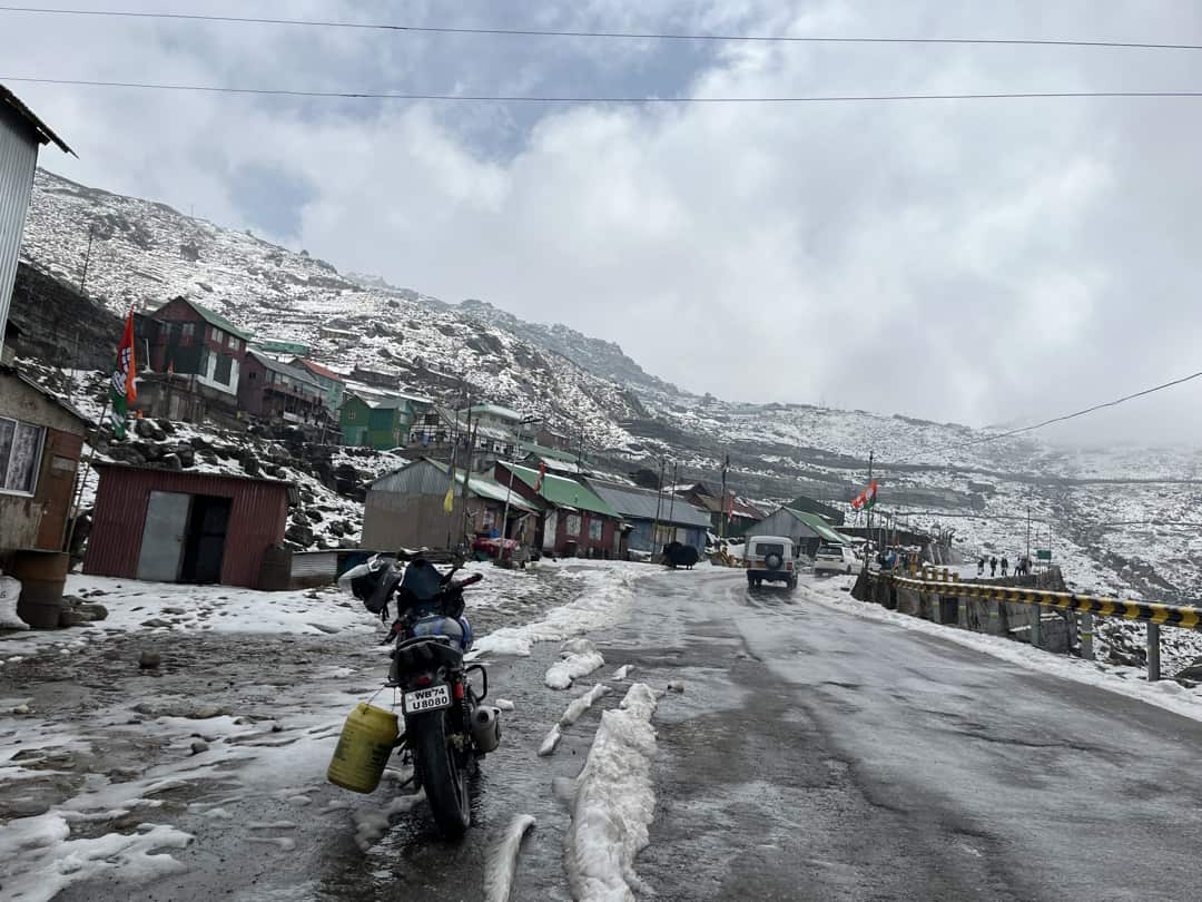 Snowy mountain village with colorful houses along a slushy road; a motorcycle parked with a yellow container nearby.