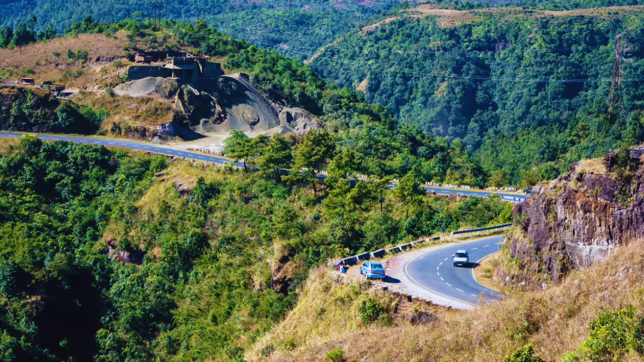 Winding mountain road with a sharp hairpin turn, guardrails, and two cars amid green hills and rocky cliffs.