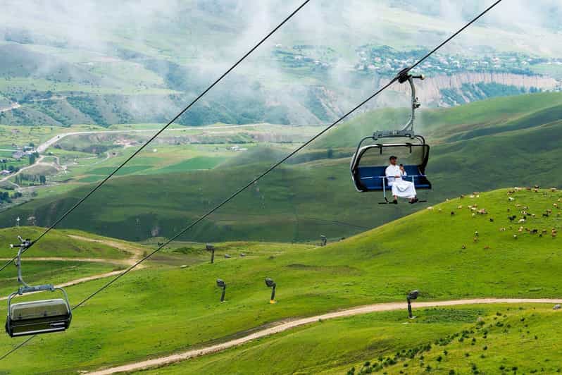 Two people ride a blue chairlift over green rolling hills with a winding dirt path and grazing animals nearby.