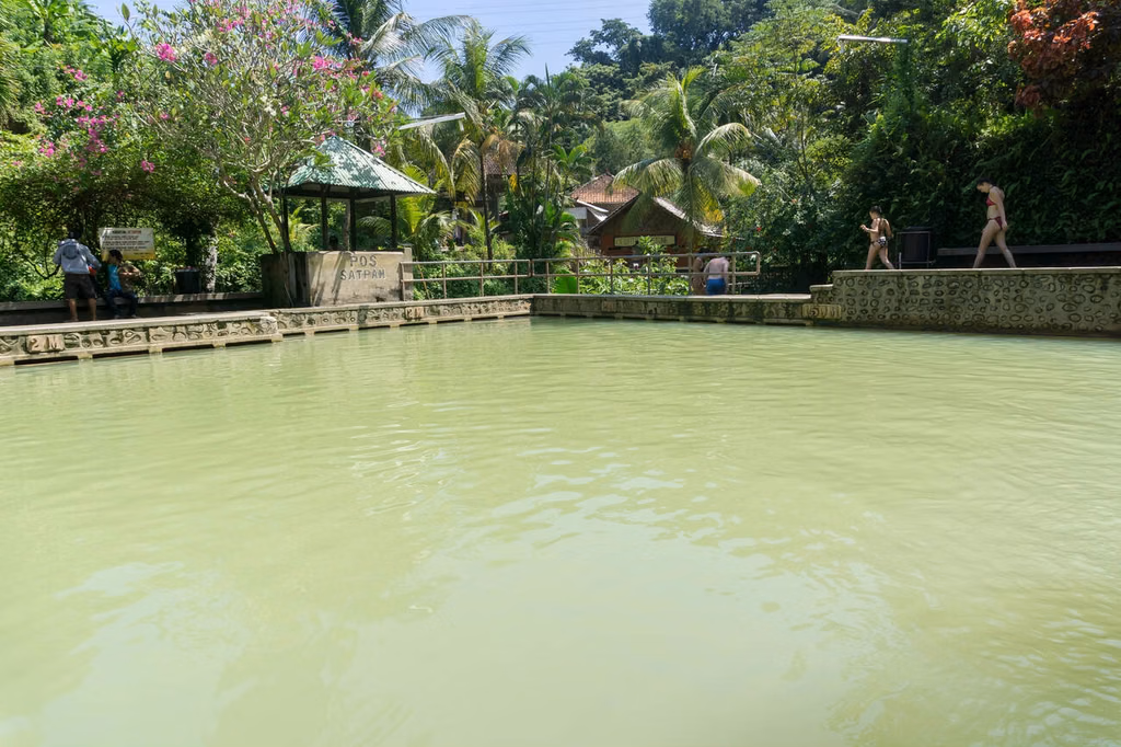 A shallow, greenish pool bordered by a stone wall and tropical trees; people in swimsuits relax along the edge under palm trees.