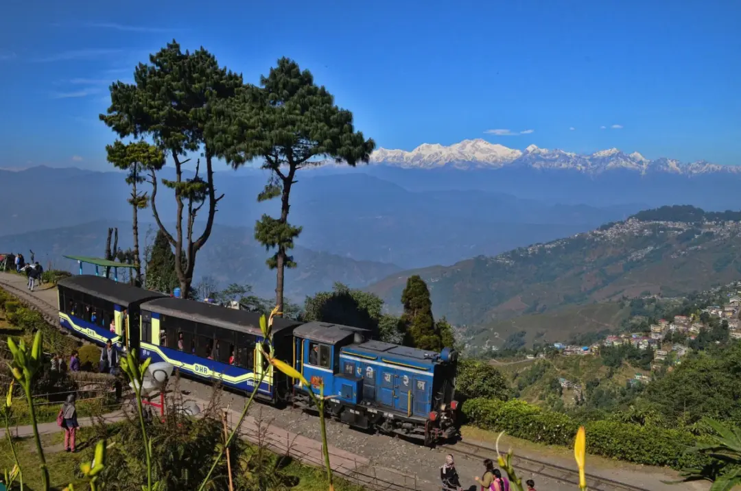 Blue tourist train on a mountain railway with snow-capped peaks in the distance and visitors on the hillside trail.