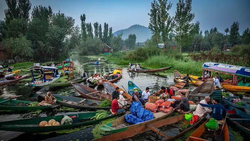 People in wooden boats on a crowded river floating market with vendors trading produce along a tree-lined bank.