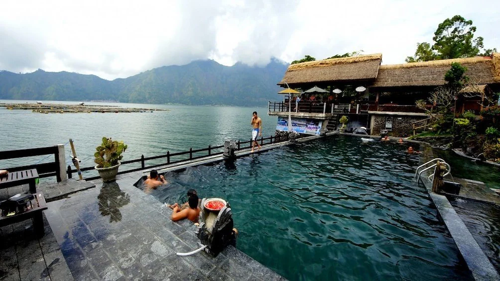 People bathing in an outdoor pool with a resort building and lake, mountains in the background