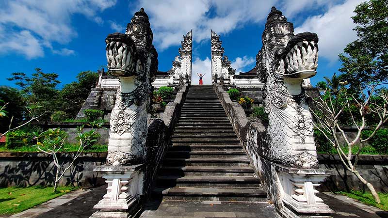 Stone staircase flanked by dragon statues leading to a temple entrance under a blue sky and green garden on both sides.