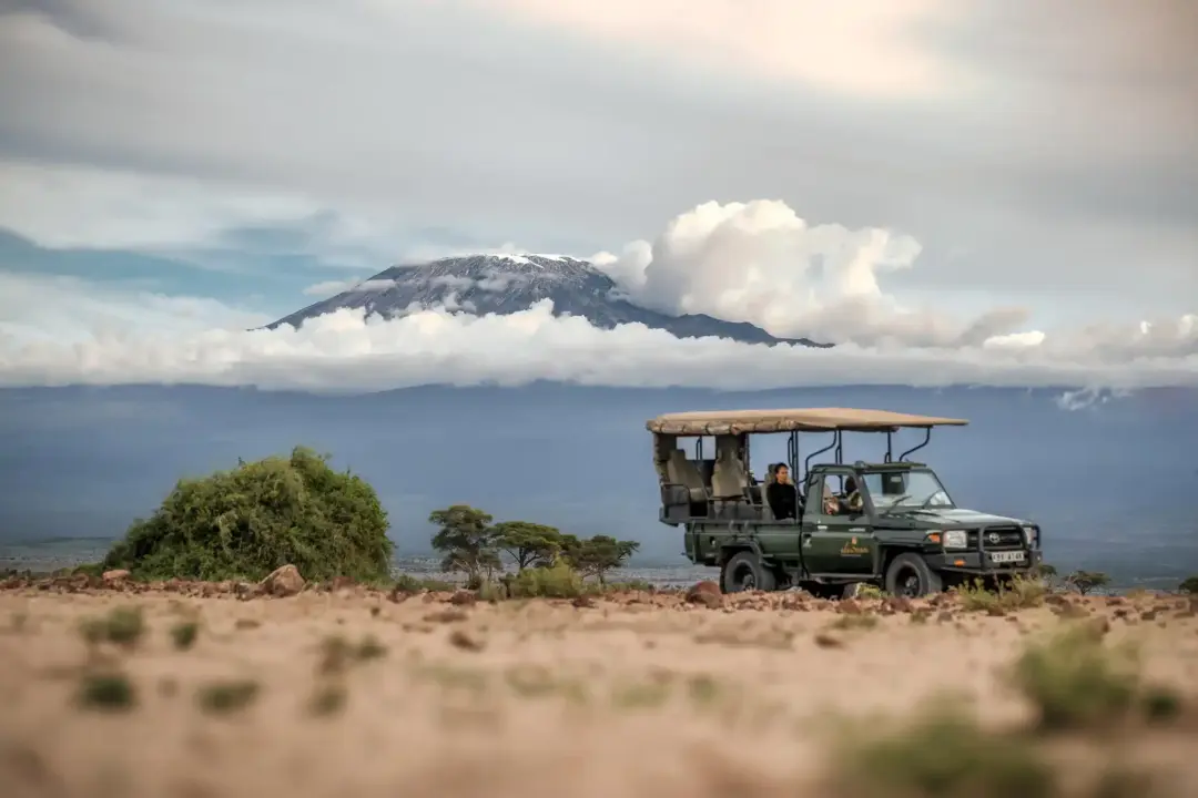 Open-roof safari jeep carrying passengers on a rocky savannah with a snow-capped mountain and clouds in the background.