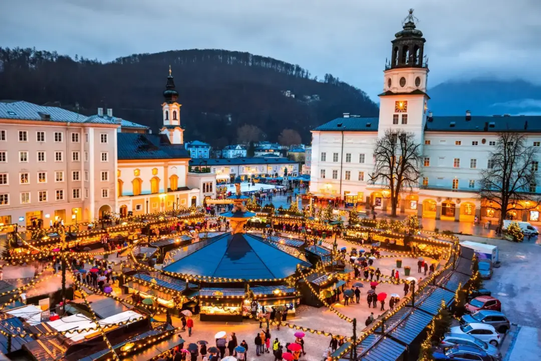 Christmas market in a town square at dusk, string-lit stalls around a central pavilion and white historic buildings with towers.
