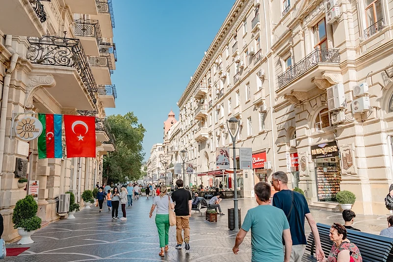 Busy sunlit street with ornate beige buildings; Azerbaijan and Turkey flags hang from a storefront on the left as pedestrians stroll past outdoor cafes.