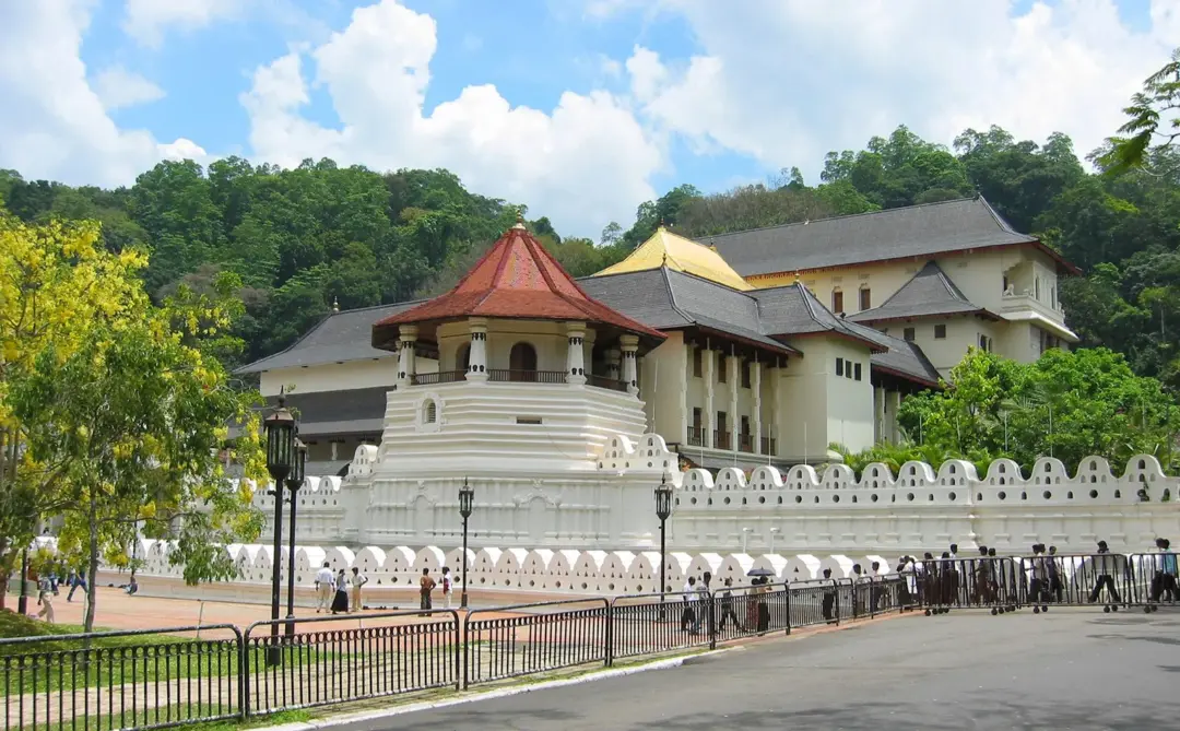 White walled temple complex with a red-roofed pavilion on a tiered base; yellow-roofed buildings sit beyond, set against green hills and a blue sky. Visitors walk along the fence.