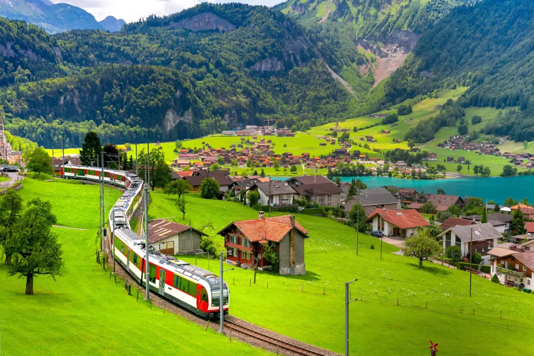 A passenger train winds along a curved track through a green valley toward a lakeside village with alpine mountains in the background.