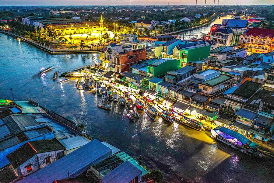 Aerial view of a busy riverside market with colorful buildings, boats docked along the water, and bright lights at dusk/dawn surrounding the scene.”,