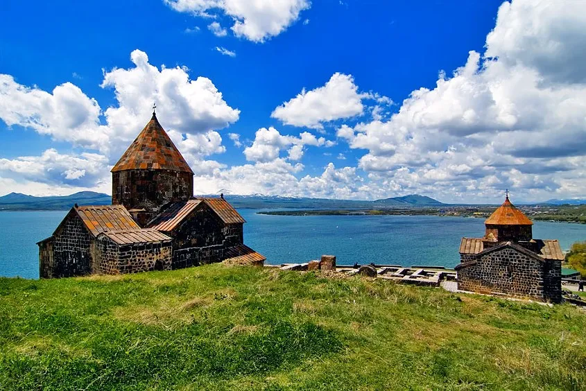 Stone fortress with two orange-tiled conical roofs on a grassy hill overlooking a blue lake and cloudy sky.