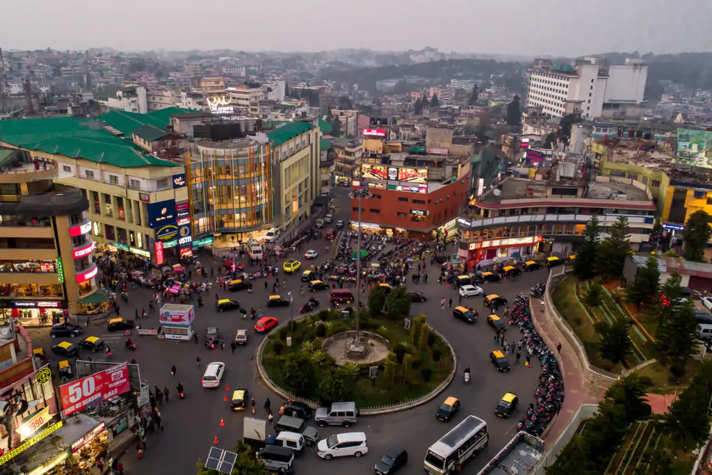 Aerial view of a busy city roundabout with cars and buses circling a green central island, surrounded by shops and bright storefronts.