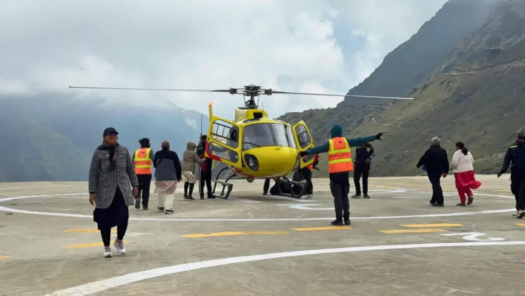 Yellow helicopter on a circular helipad amid group of people in jackets, with alpine mountains in the background and some crew in orange vests guiding it