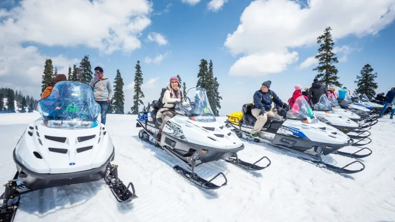Group of people in winter gear standing near a row of white snowmobiles on a snowy field with trees and blue sky inspired by a sunny day','Riders sit on snowmobiles, with trees and blue sky in a mountain setting','Line of snowmobiles ready for a ride in a snowy landscape with evergreen trees','People preparing for a snowmobile tour on a clear winter day','Snowmobile gathering on a snowy plain under a bright blue sky