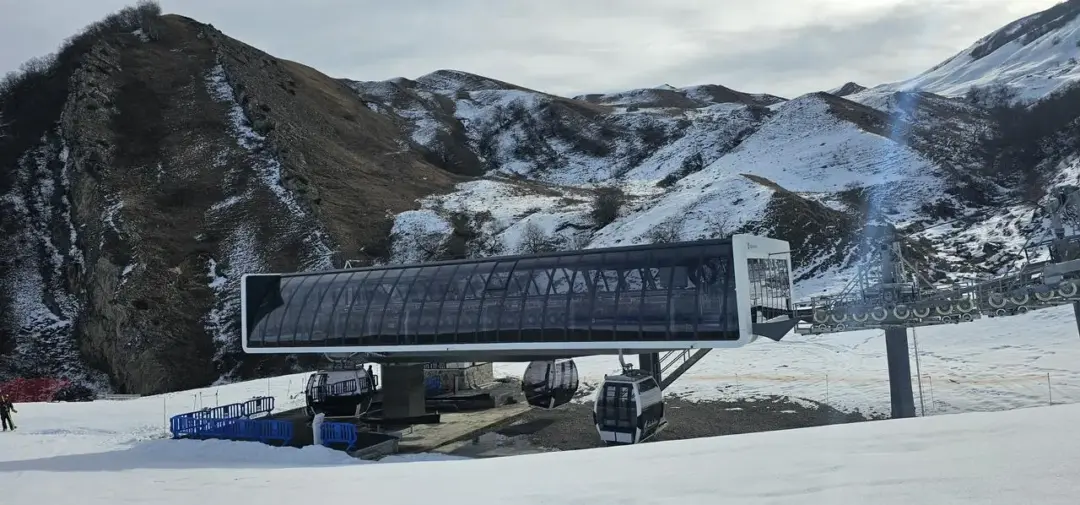 Ski lift station with glass-walled cabins on a platform, set against snow-covered mountains and slopes.